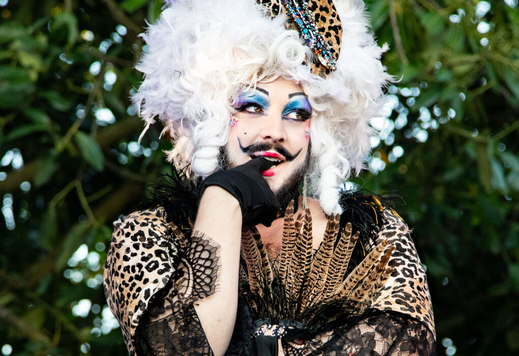Portrait de l'artiste drag Melba Zigomar en costume animalier et dentelle noire, coiffée d’une perruque blanche volumineuse décorée de plumes et motifs léopard, posant en extérieur.