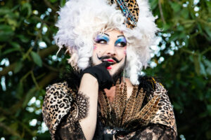 Portrait de l'artiste drag Melba Zigomar en costume animalier et dentelle noire, coiffée d’une perruque blanche volumineuse décorée de plumes et motifs léopard, posant en extérieur.
