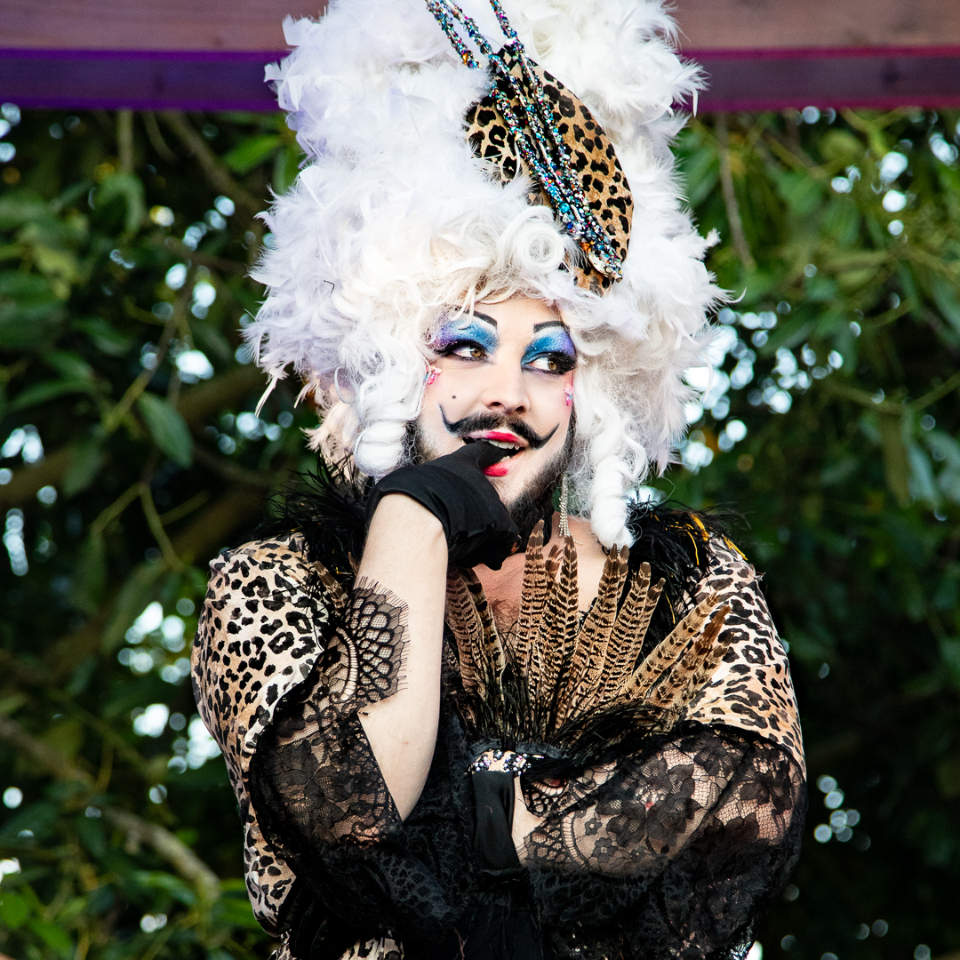 Portrait de l'artiste drag Melba Zigomar en costume animalier et dentelle noire, coiffée d’une perruque blanche volumineuse décorée de plumes et motifs léopard, posant en extérieur.