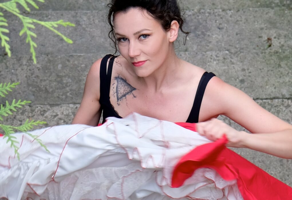 Portrait d’Andrée Gine, artiste burlesque, à l’extérieur, tenant une jupe à volants rouges et blancs, bras levés, visage partiellement tourné vers la gauche.