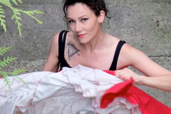 Portrait d’Andrée Gine, artiste burlesque, à l’extérieur, tenant une jupe à volants rouges et blancs, bras levés, visage partiellement tourné vers la gauche.