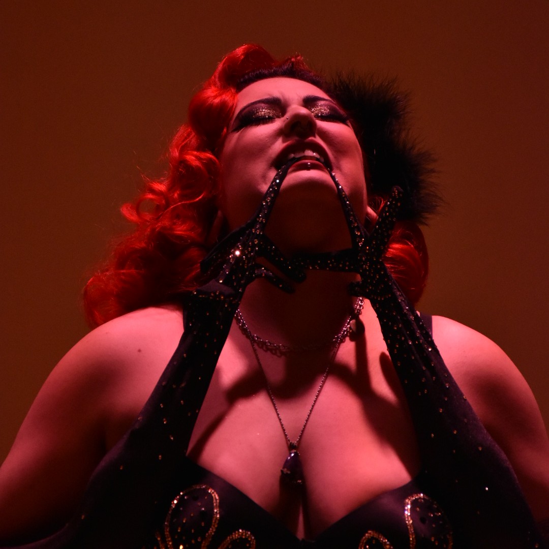 Gros plan de Scarlett Von Rubis, chevelure rouge ondulée, en lingerie noire pailletée et gants longs, posant la tête renversée en arrière dans une lumière rouge.