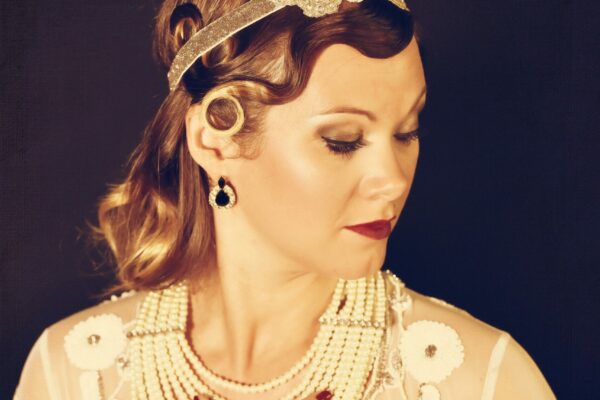 Portrait glamour de Delis MissKitty en style années 20, portant un bandeau bijouté, un collier de perles superposé et une veste transparente brodée, main posée sur la poitrine.