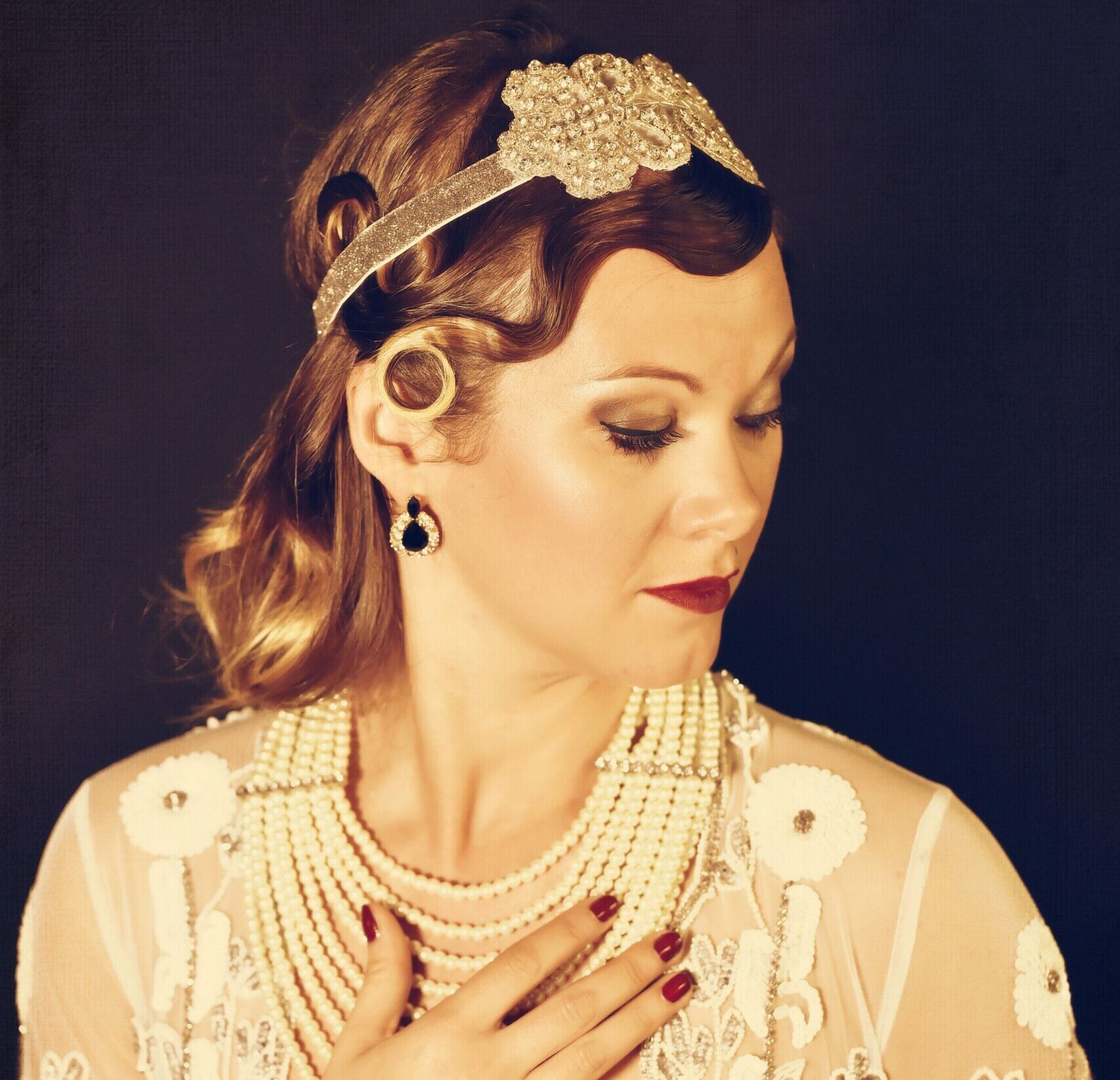 Portrait glamour de Delis MissKitty en style années 20, portant un bandeau bijouté, un collier de perles superposé et une veste transparente brodée, main posée sur la poitrine.