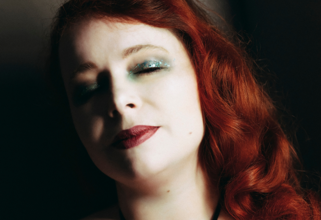 Portrait rapproché de Julie Crève-Coeur mettant en valeur ses cheveux roux ondulés et son décolleté dans un costume de scène noir.