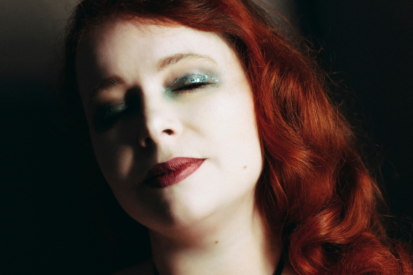 Portrait rapproché de Julie Crève-Coeur mettant en valeur ses cheveux roux ondulés et son décolleté dans un costume de scène noir.