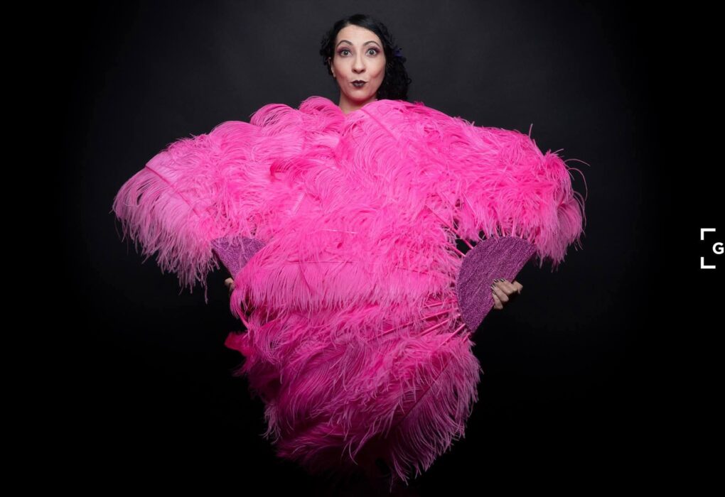 Portrait d’Eva La Vamp de dos, enveloppée dans un grand boa d’éventails roses sur fond noir, photographiée en studio.