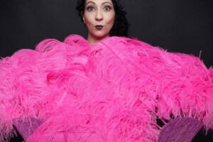Portrait d’Eva La Vamp de dos, enveloppée dans un grand boa d’éventails roses sur fond noir, photographiée en studio.