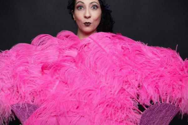 Portrait d’Eva La Vamp de dos, enveloppée dans un grand boa d’éventails roses sur fond noir, photographiée en studio.