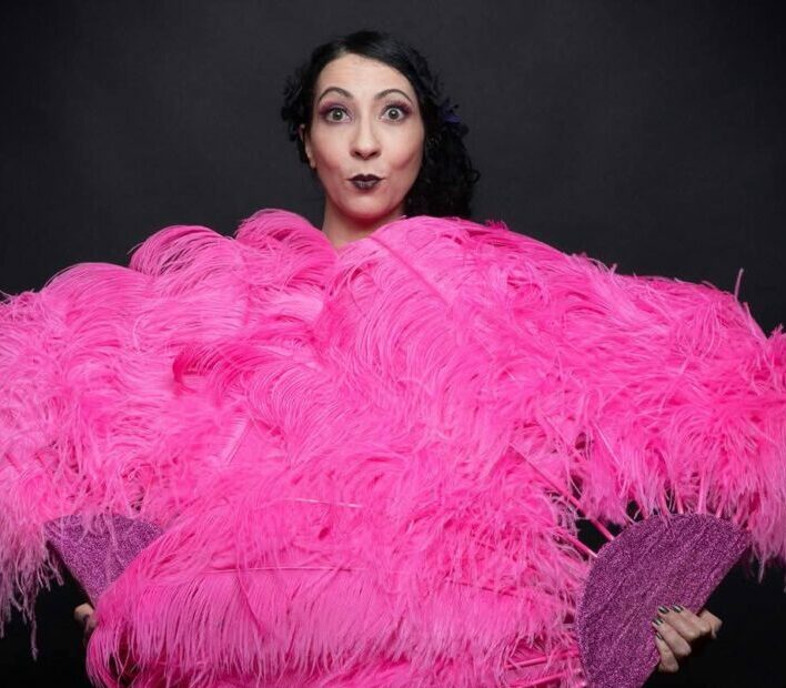 Portrait d’Eva La Vamp de dos, enveloppée dans un grand boa d’éventails roses sur fond noir, photographiée en studio.