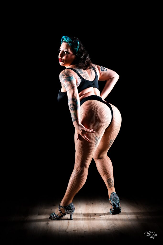 Vue de dos en pied de l’artiste burlesque Gemma Lefik en lingerie noire, résille couleur chair et chaussures à talons scintillantes, posant sur un parquet éclairé devant un fond noir.