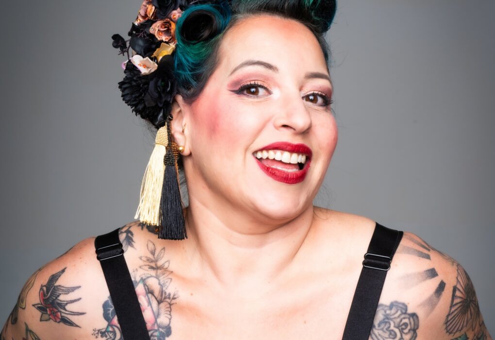 Portrait studio de l’artiste burlesque Gemma Lefik souriante, en tenue pin-up imprimée, coiffure rétro avec fleurs noires et dorées et maquillage scénique marqué, sur fond gris uni.