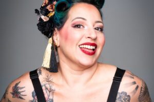 Portrait studio de l’artiste burlesque Gemma Lefik souriante, en tenue pin-up imprimée, coiffure rétro avec fleurs noires et dorées et maquillage scénique marqué, sur fond gris uni.