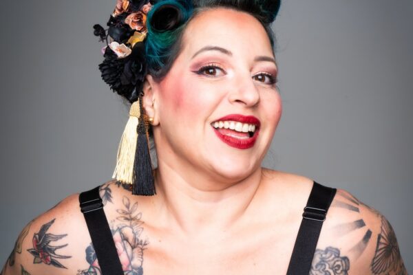 Portrait studio de l’artiste burlesque Gemma Lefik souriante, en tenue pin-up imprimée, coiffure rétro avec fleurs noires et dorées et maquillage scénique marqué, sur fond gris uni.
