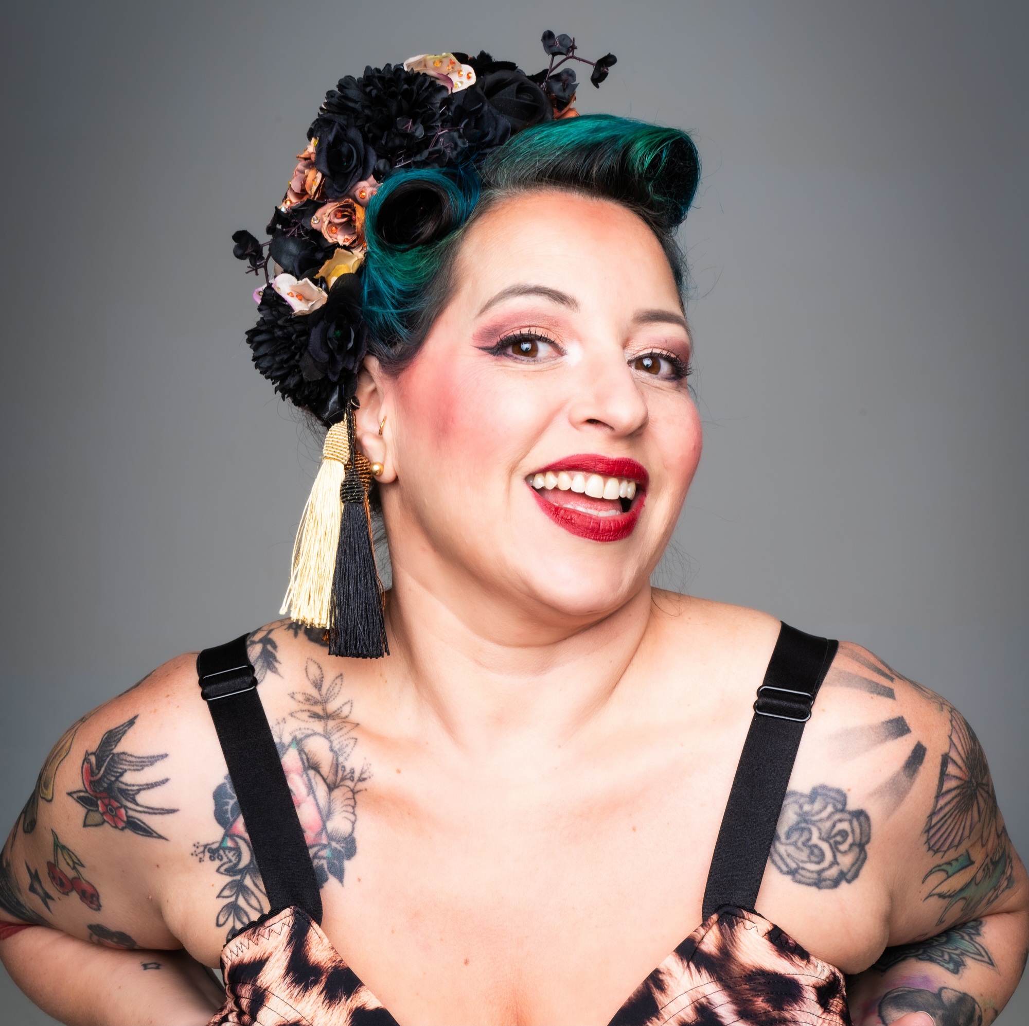 Portrait studio de l’artiste burlesque Gemma Lefik souriante, en tenue pin-up imprimée, coiffure rétro avec fleurs noires et dorées et maquillage scénique marqué, sur fond gris uni.