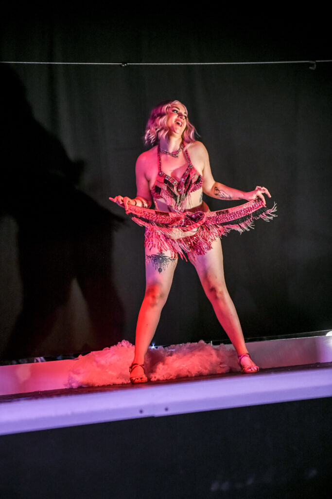 Artiste burlesque Stella Luna en costume à franges roses, souriante sur scène, tenant sa jupe durant une performance, photographiée par ericl14.el.