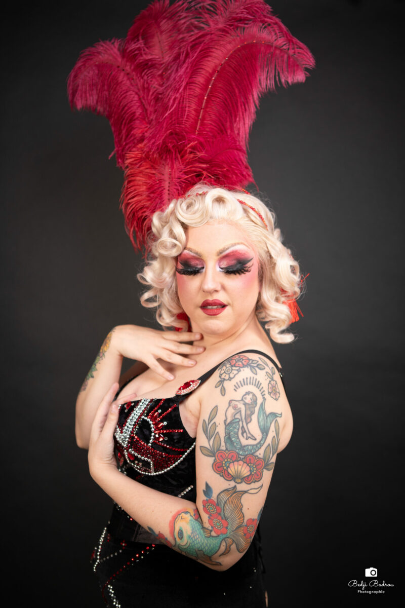 Betty Be Good en portrait burlesque glamour, grande coiffe à plumes rouges, bustier noir orné de sirène tatouée, maquillage dramatique – Photo Bedji Badrou
