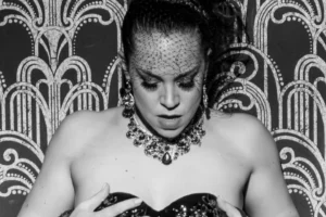 Portrait noir et blanc de l’artiste burlesque Lulu Madeleine, de face, tenant son bustier décoré avec les mains devant un fond graphique art déco, photographiée par Christian Kieffer.