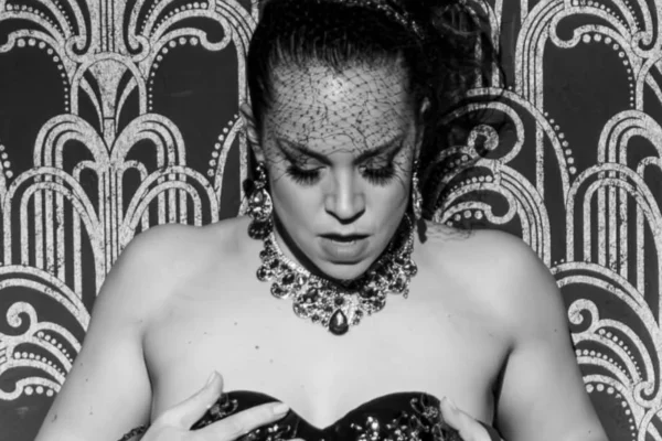Portrait noir et blanc de l’artiste burlesque Lulu Madeleine, de face, tenant son bustier décoré avec les mains devant un fond graphique art déco, photographiée par Christian Kieffer.