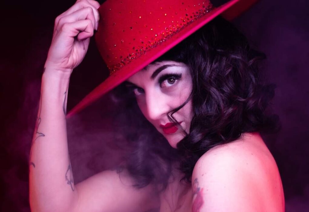 Madâme Plum'ti en portrait burlesque, chapeau rouge à large bord, lumière rose et fumée de scène