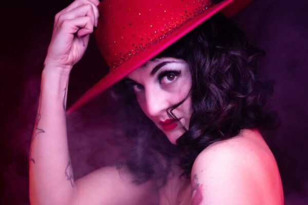 Madâme Plum'ti en portrait burlesque, chapeau rouge à large bord, lumière rose et fumée de scène