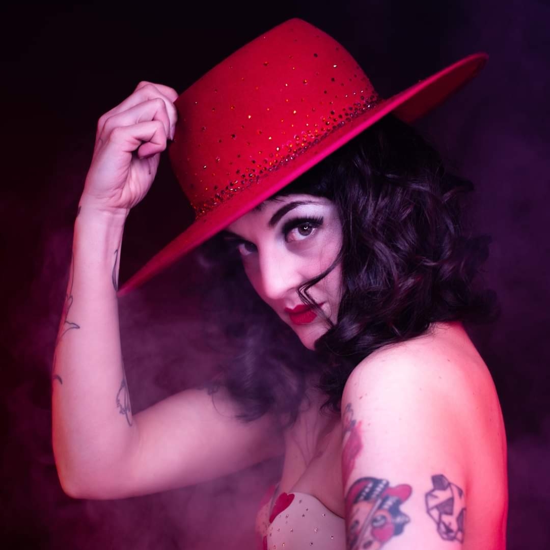 Madâme Plum'ti en portrait burlesque, chapeau rouge à large bord, lumière rose et fumée de scène