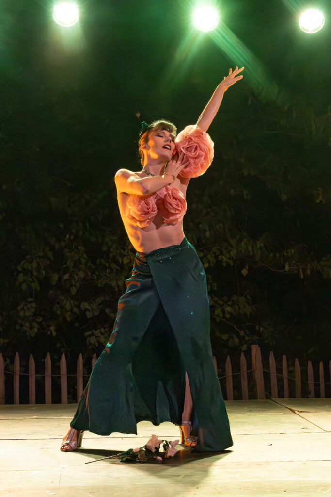 Andrée Gine sur scène, bras levés, vêtue d’une jupe longue et d’une brassière ornée de grandes fleurs roses sous des projecteurs, décor extérieur nocturne.