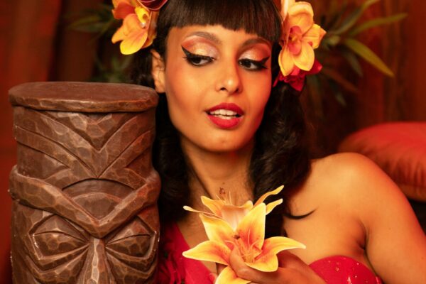 Portrait de Froufrou d’Absinthe en tenue rouge tropicale, coiffée de fleurs jaunes et rouges, posant à côté d’une grande statue tiki sculptée dans une ambiance chaleureuse.