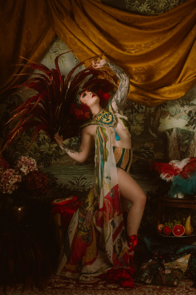 Kiki Béguin sur scène en costume burlesque baroque, tenant un éventail de plumes rouges, devant un décor de drapés dorés et de nature peinte, photographiée par Emilia Lyon.