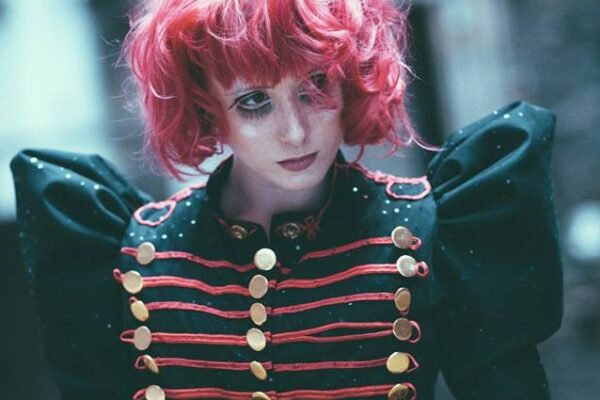 Portrait de l’artiste burlesque Kiki Béguin aux cheveux roses, vêtue d’une veste noire militaire ornée de galons rouges, photographiée par Sophie Alyz.