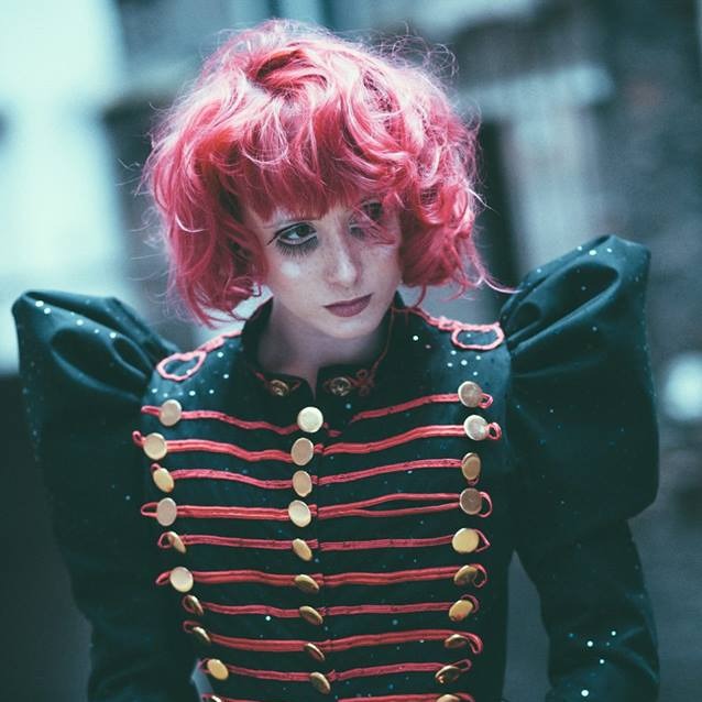 Portrait de l’artiste burlesque Kiki Béguin aux cheveux roses, vêtue d’une veste noire militaire ornée de galons rouges, photographiée par Sophie Alyz.