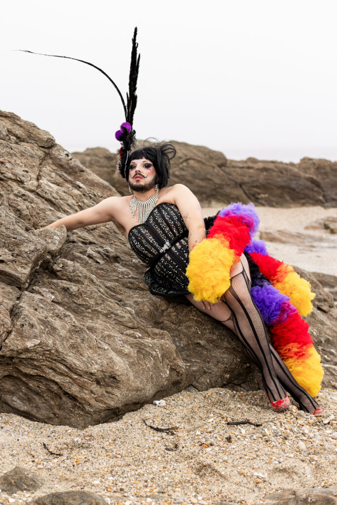 Melba Zigomar allongée sur un rocher en bord de mer, portant une robe noire à strass, collants résille et boa multicolore, coiffée d’un grand headpiece à plumes noires.