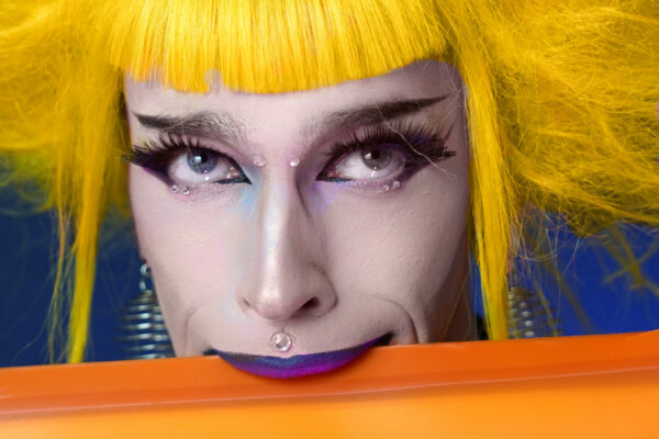 Portrait serré de l’artiste burlesque Mélusine Beauséjour, coiffée d’une perruque jaune volumineuse, derrière une surface orange, photographiée par Alexia Rince.
