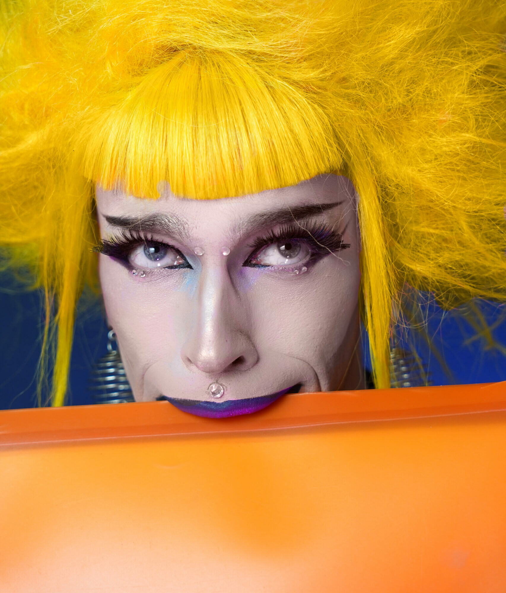 Portrait serré de l’artiste burlesque Mélusine Beauséjour, coiffée d’une perruque jaune volumineuse, derrière une surface orange, photographiée par Alexia Rince.