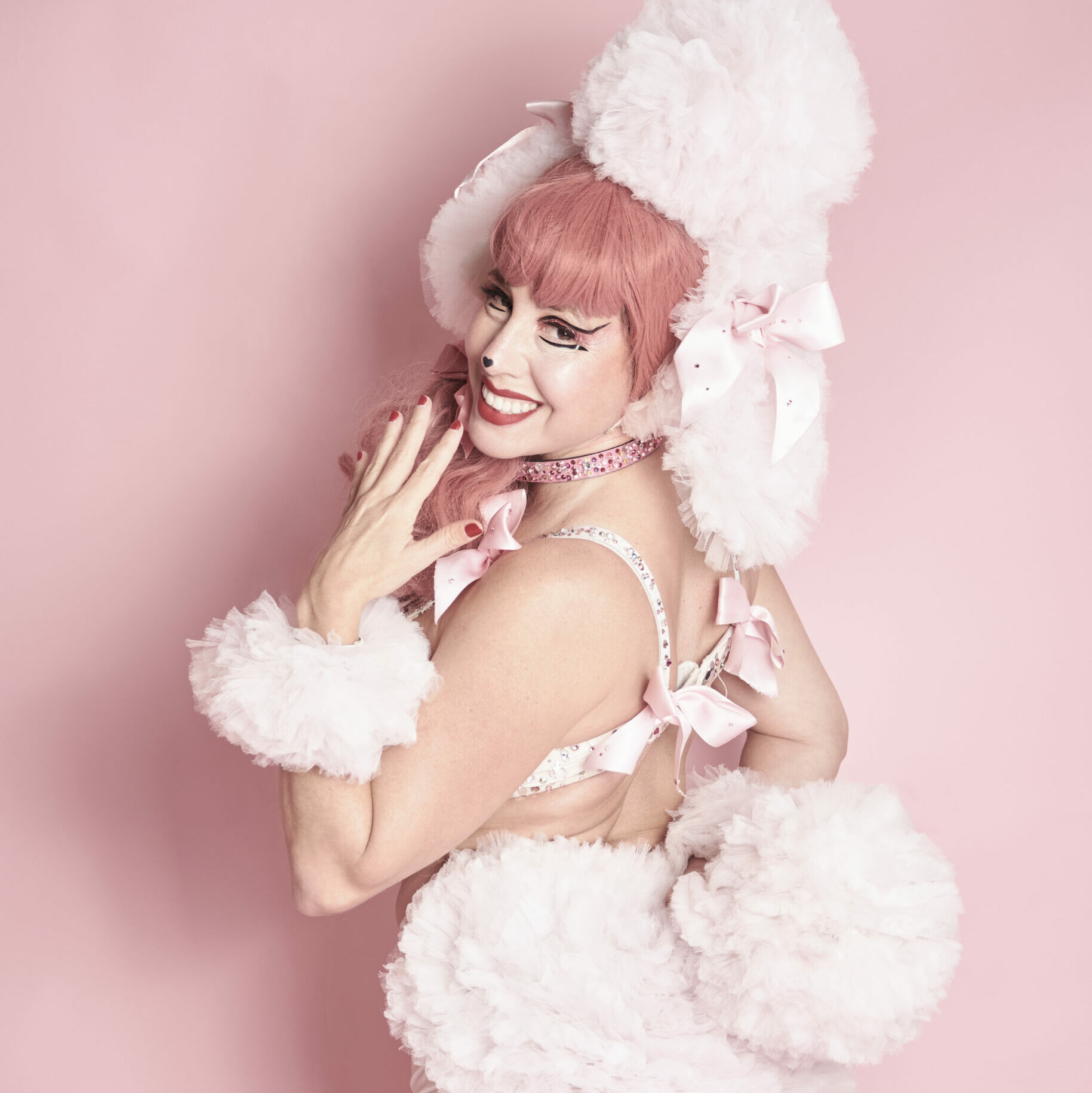 Rosabelle Sélavy de dos en costume burlesque rose et blanc inspiré d’un caniche, posant devant un fond rose en studio, photographiée par Studio Volua à Paris.