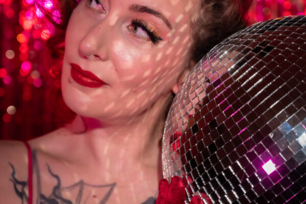 Nasty Von Trier en gros plan, en robe à paillettes rouges et gants assortis, tenant une boule à facettes devant un fond scintillant rose, photographiée par Gilles Baermann.