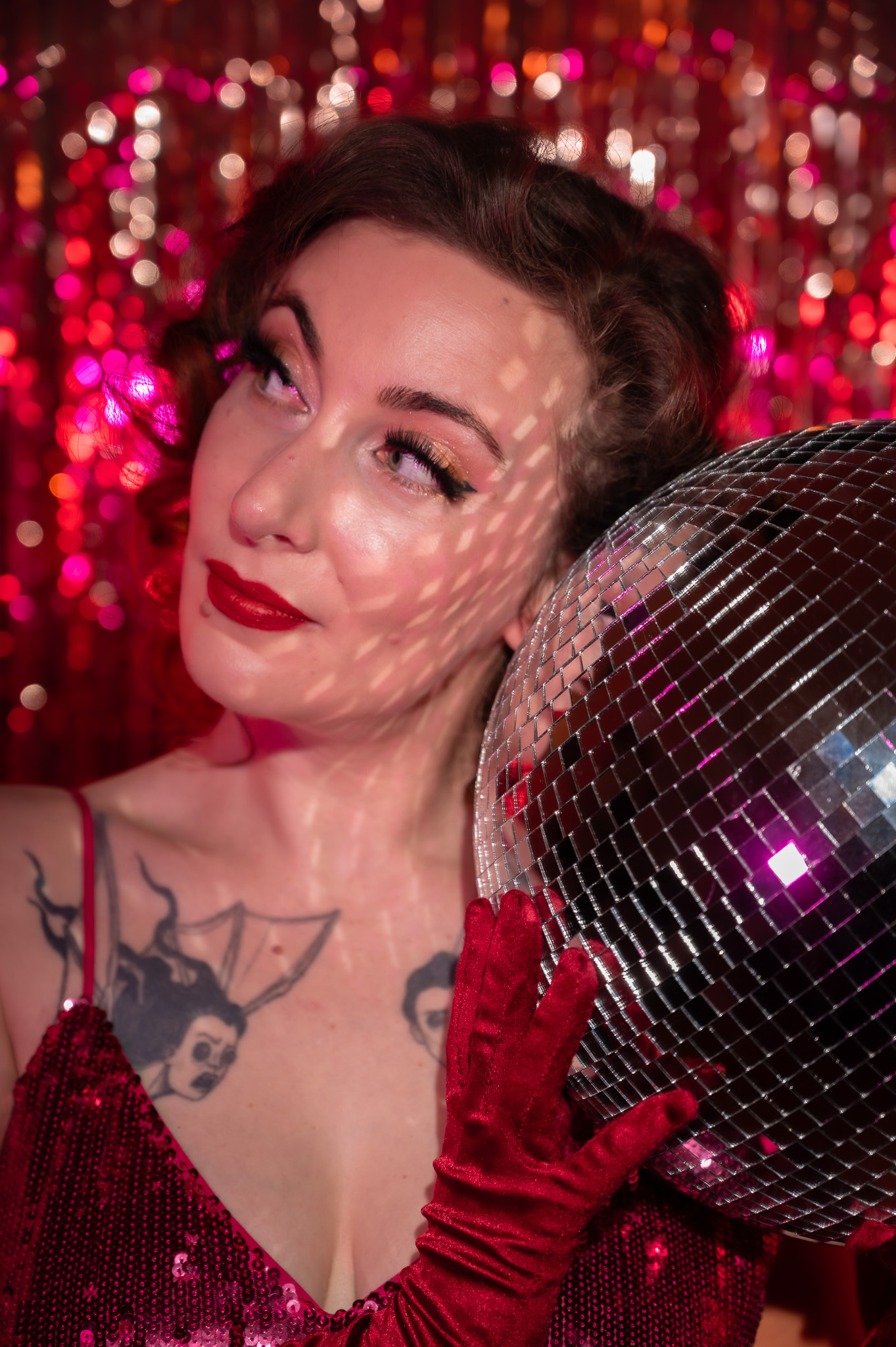 Nasty Von Trier en gros plan, en robe à paillettes rouges et gants assortis, tenant une boule à facettes devant un fond scintillant rose, photographiée par Gilles Baermann.