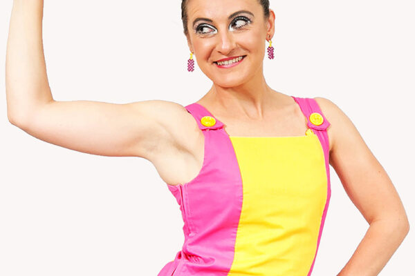 Plan rapproché de Coney Bow en robe bicolore rose et jaune, tenant sa queue de cheval haute d’une main, sur fond clair, avec accessoires colorés.