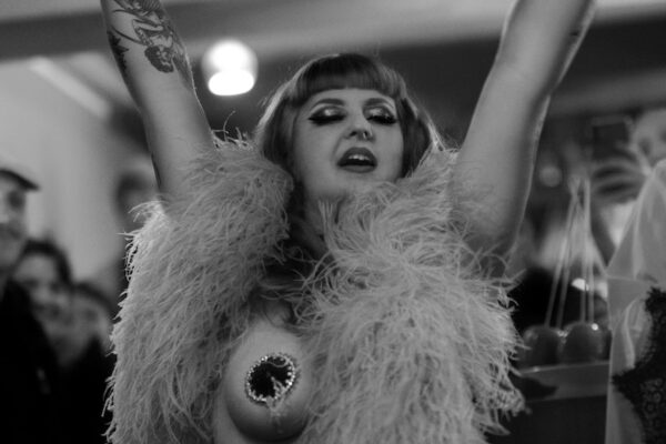 Maureen Blue Lagoon en performance burlesque, portrait en noir et blanc, bras levés, portant un boa à plumes, devant un public en arrière-plan – Photo Thibault Pierrisnard.