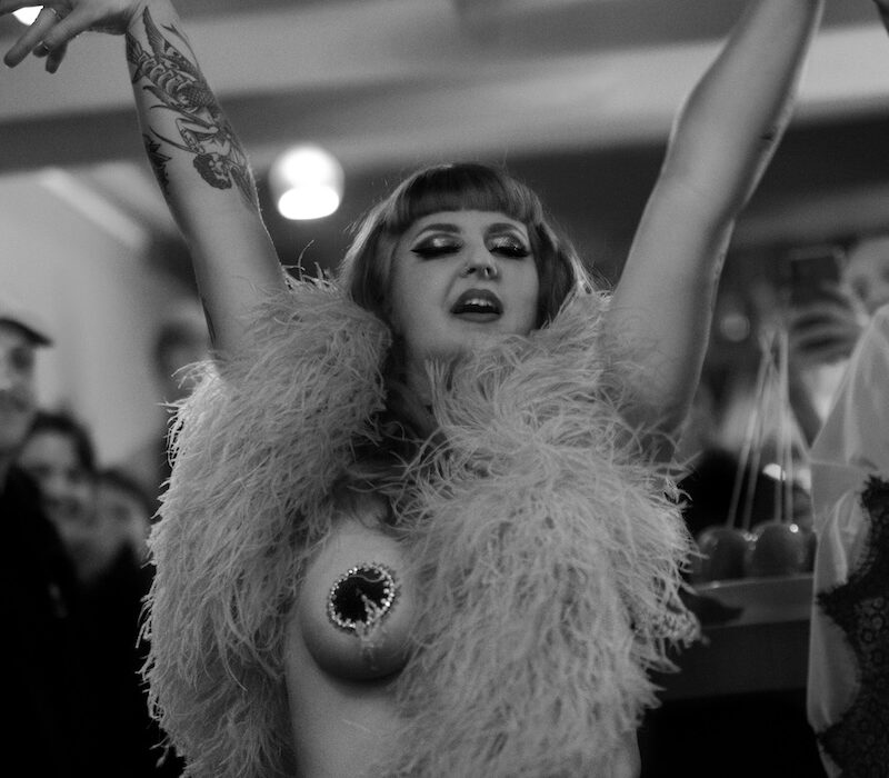 Maureen Blue Lagoon en performance burlesque, portrait en noir et blanc, bras levés, portant un boa à plumes, devant un public en arrière-plan – Photo Thibault Pierrisnard.