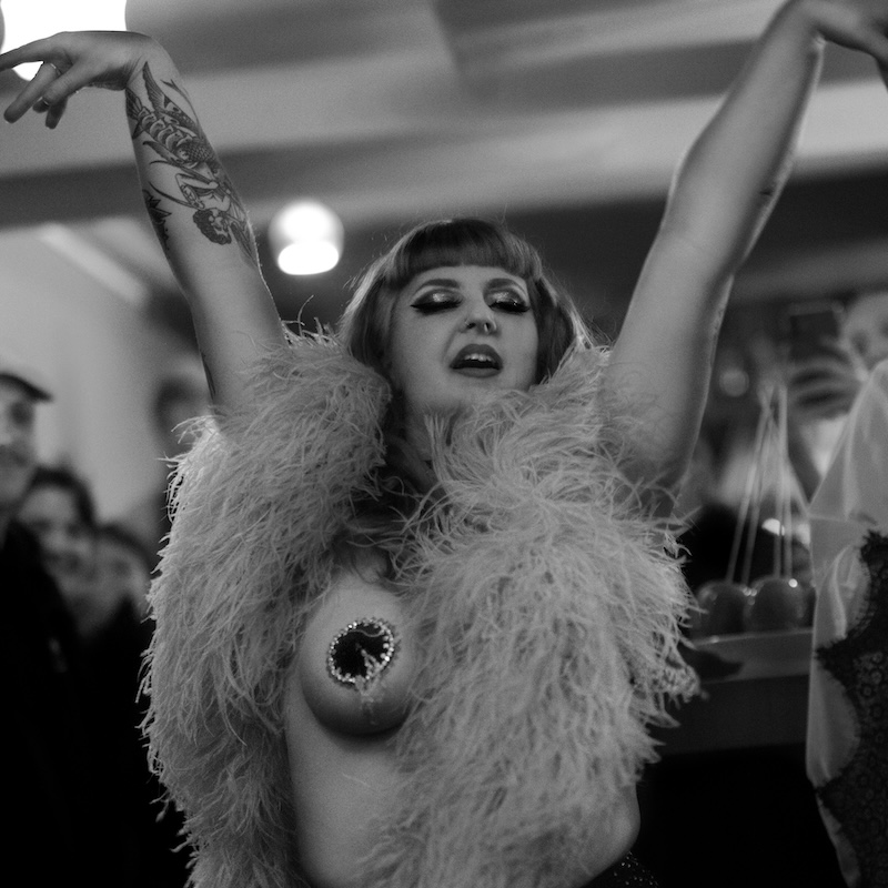 Maureen Blue Lagoon en performance burlesque, portrait en noir et blanc, bras levés, portant un boa à plumes, devant un public en arrière-plan – Photo Thibault Pierrisnard.