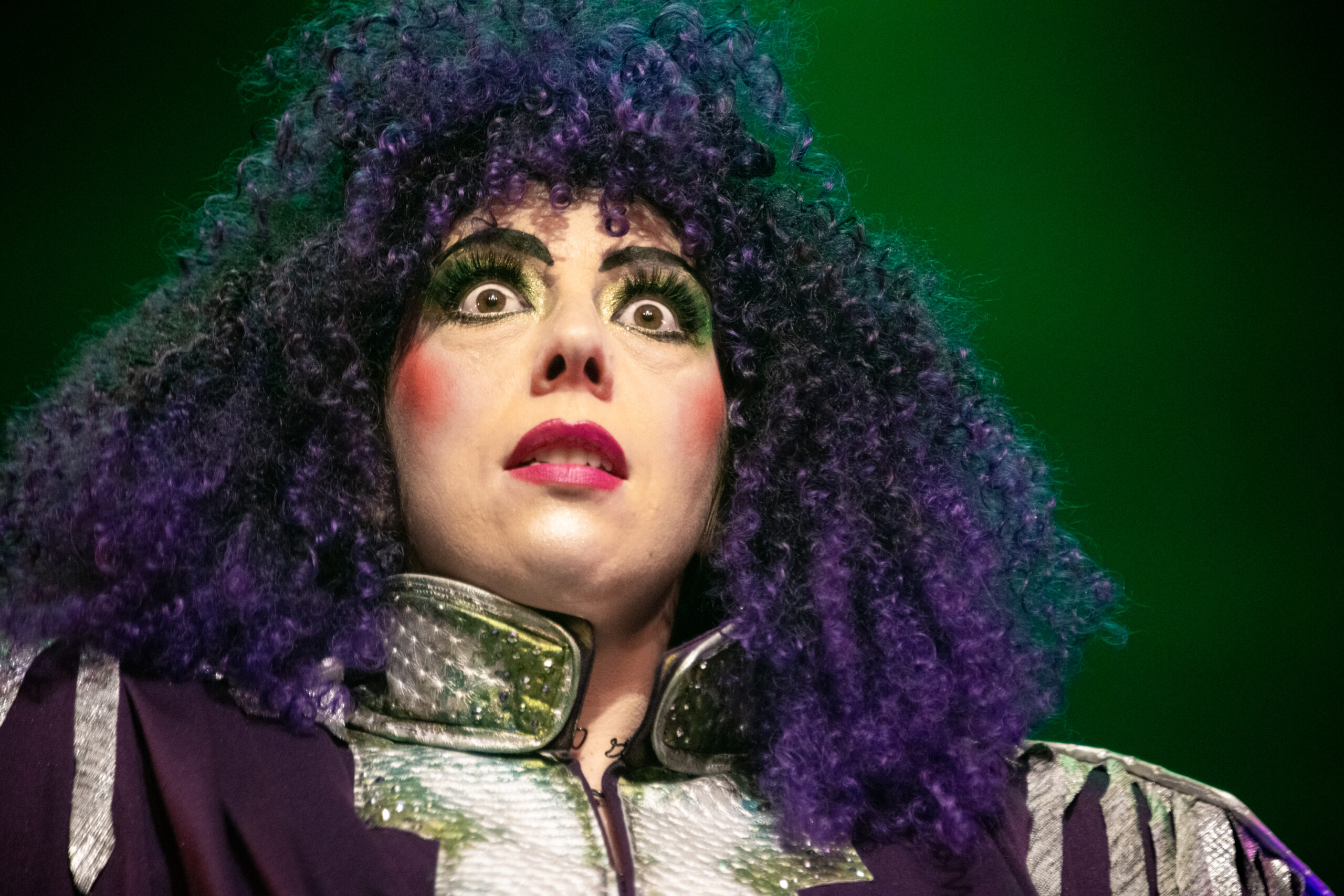 Portrait de l’artiste burlesque Pixie Vixen sur scène, perruque bouclée violette volumineuse et costume scintillant vert et argent, photographiée par Noire Chambre.