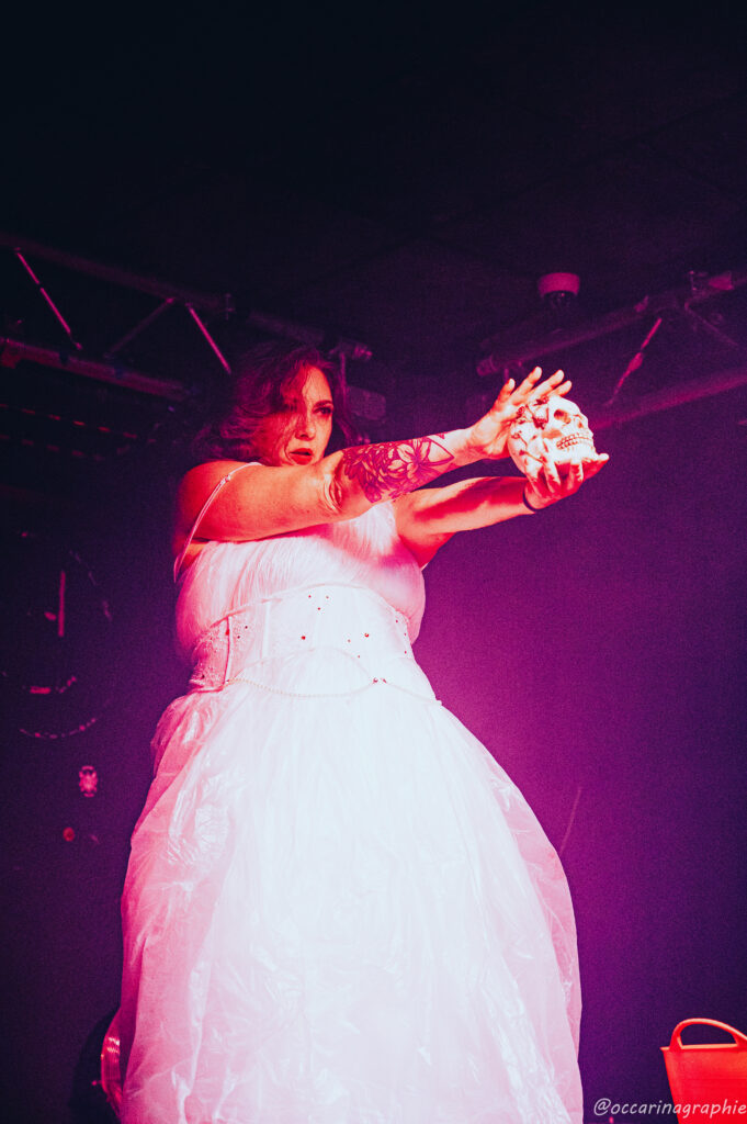 Malicia Kiss en robe blanche de mariée, tendant un crâne décoré vers le public sur scène, dans une lumière rose, photographiée par Occarinagraphie.