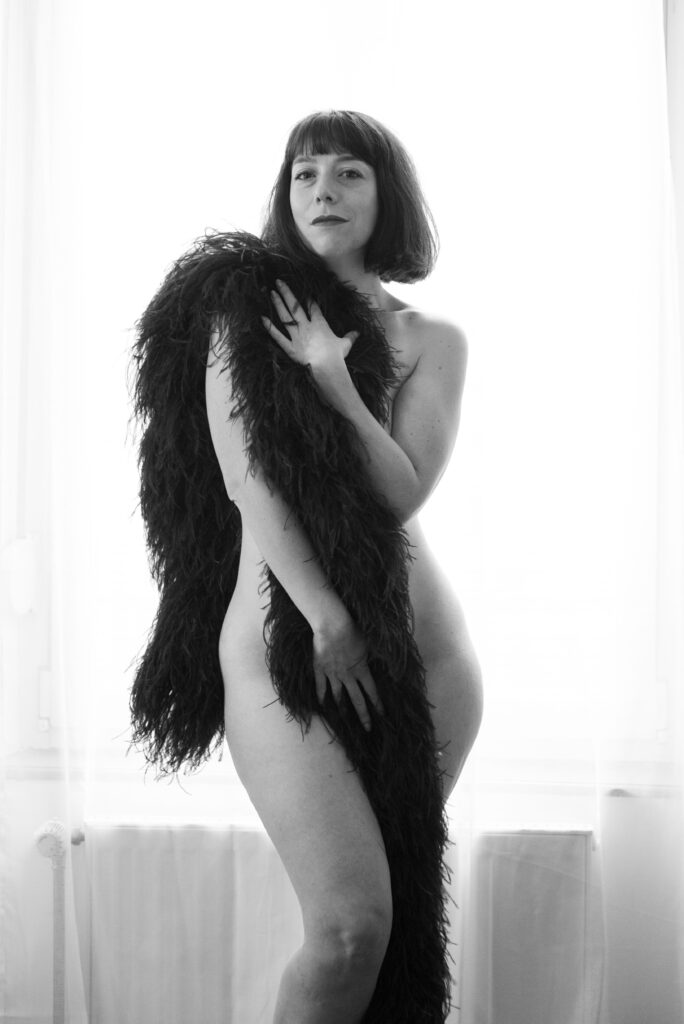 Pixie Vixen posant nue en noir et blanc, partiellement dissimulée derrière un long boa de plumes sombres, devant une fenêtre lumineuse, photographiée par Deborah Gigliotti.