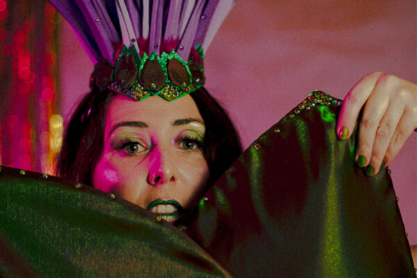 Portrait rapproché d’Artye Chaude en headdress plume violet et vert tenant son costume végétal devant le visage