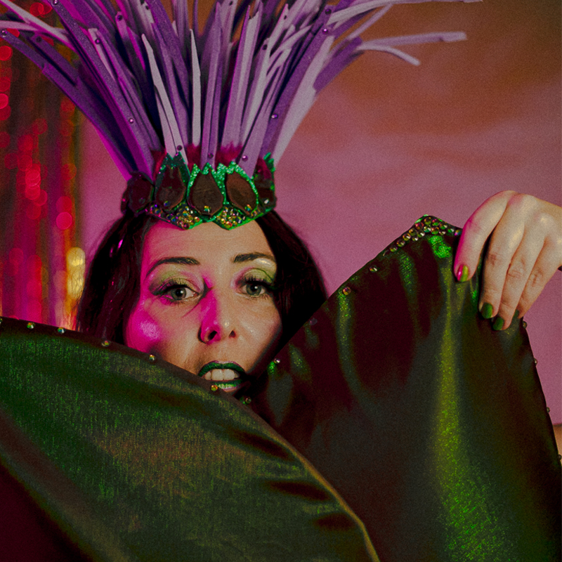 Portrait rapproché d’Artye Chaude en headdress plume violet et vert tenant son costume végétal devant le visage