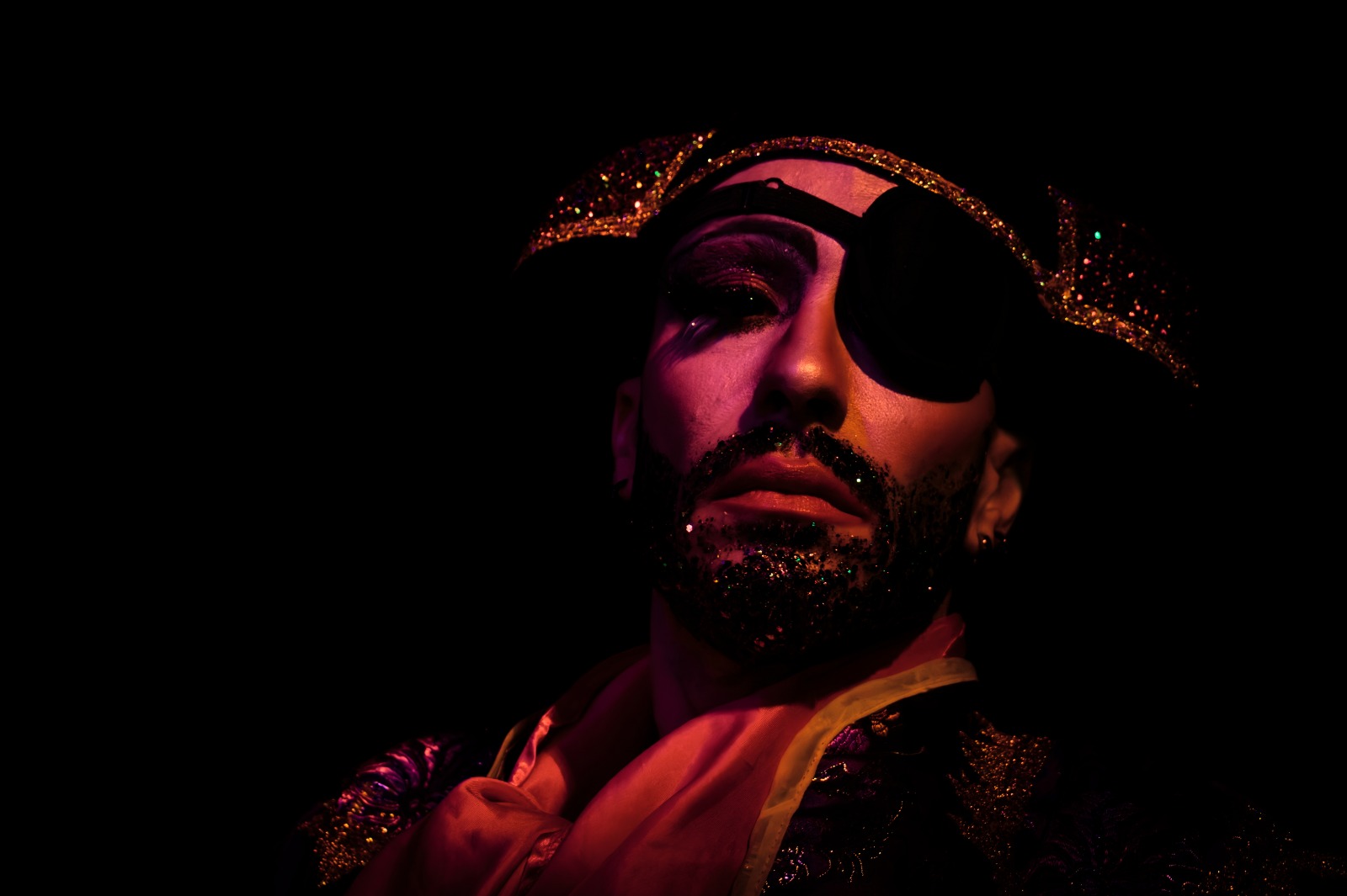 Artiste burlesque Figo-Lou en costume sombre à paillettes, partiellement éclairé sur fond noir, portant un tricorne brillant, photographié par @glhem.