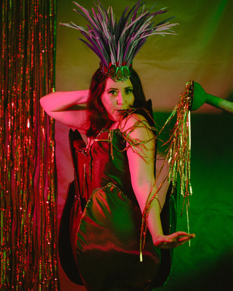 Artiste burlesque Artye Chaude en costume végétal vert et violet sur scène, sous lumières rouges et vertes