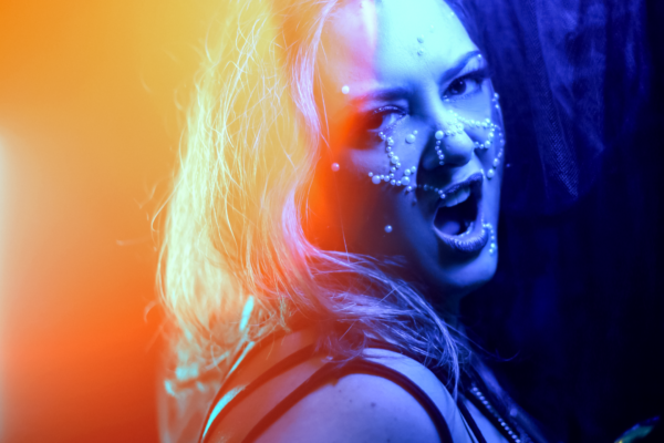 Portrait de l’artiste burlesque Malicia Kiss, visage orné de perles blanches, éclairée en bleu et orange dans une ambiance de scène, photographiée par The Fetching Eye.