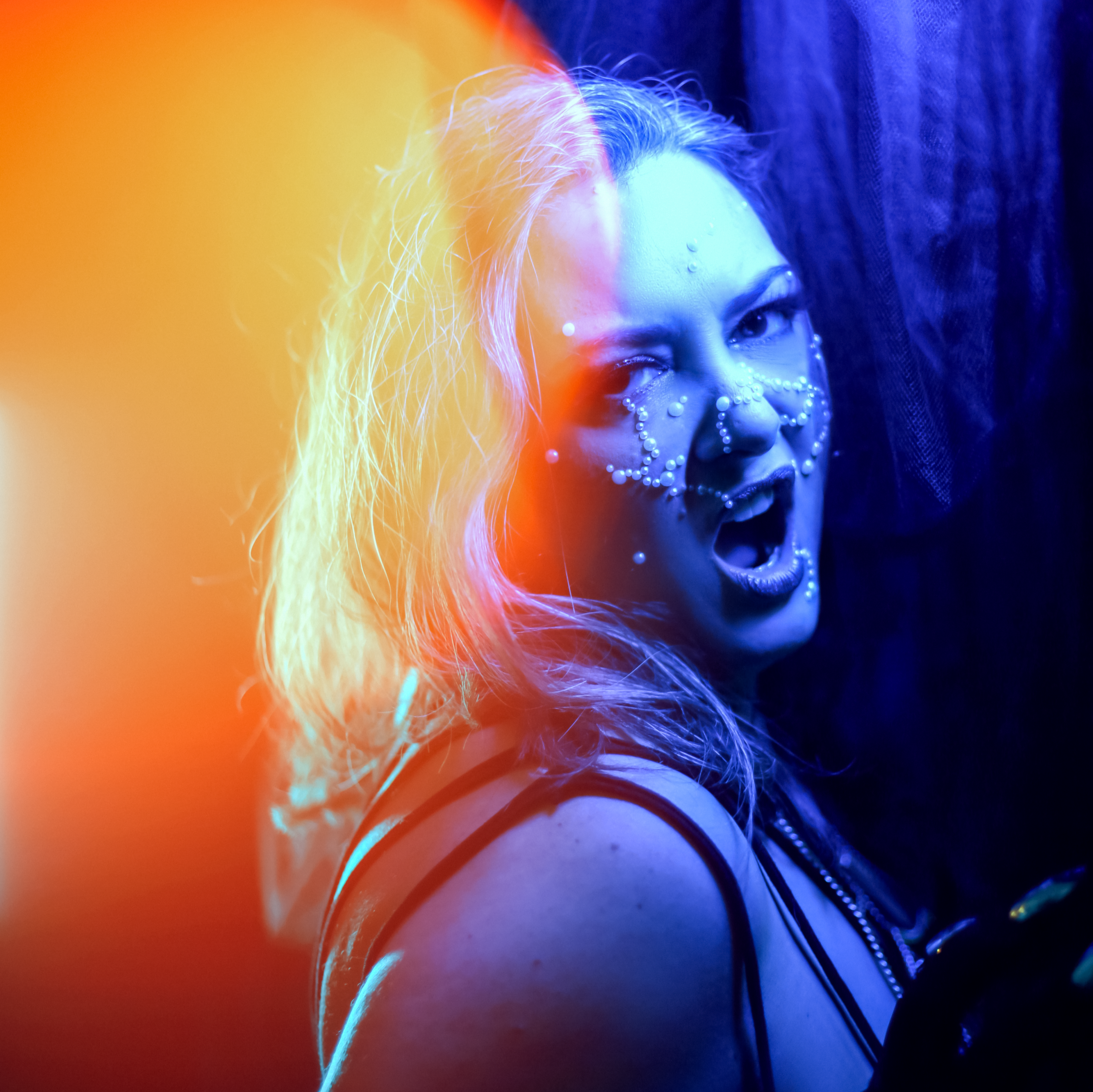 Portrait de l’artiste burlesque Malicia Kiss, visage orné de perles blanches, éclairée en bleu et orange dans une ambiance de scène, photographiée par The Fetching Eye.